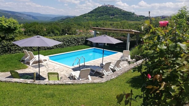 Piscina con sillones de descanso y sombrillas, rodeada de césped y con vistas a una colina con un edificio en la cima.