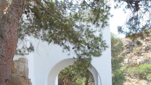 Un sendero de tierra que lleva a un edificio blanco con entrada abovedada, rodeado de árboles y terreno rocoso.
