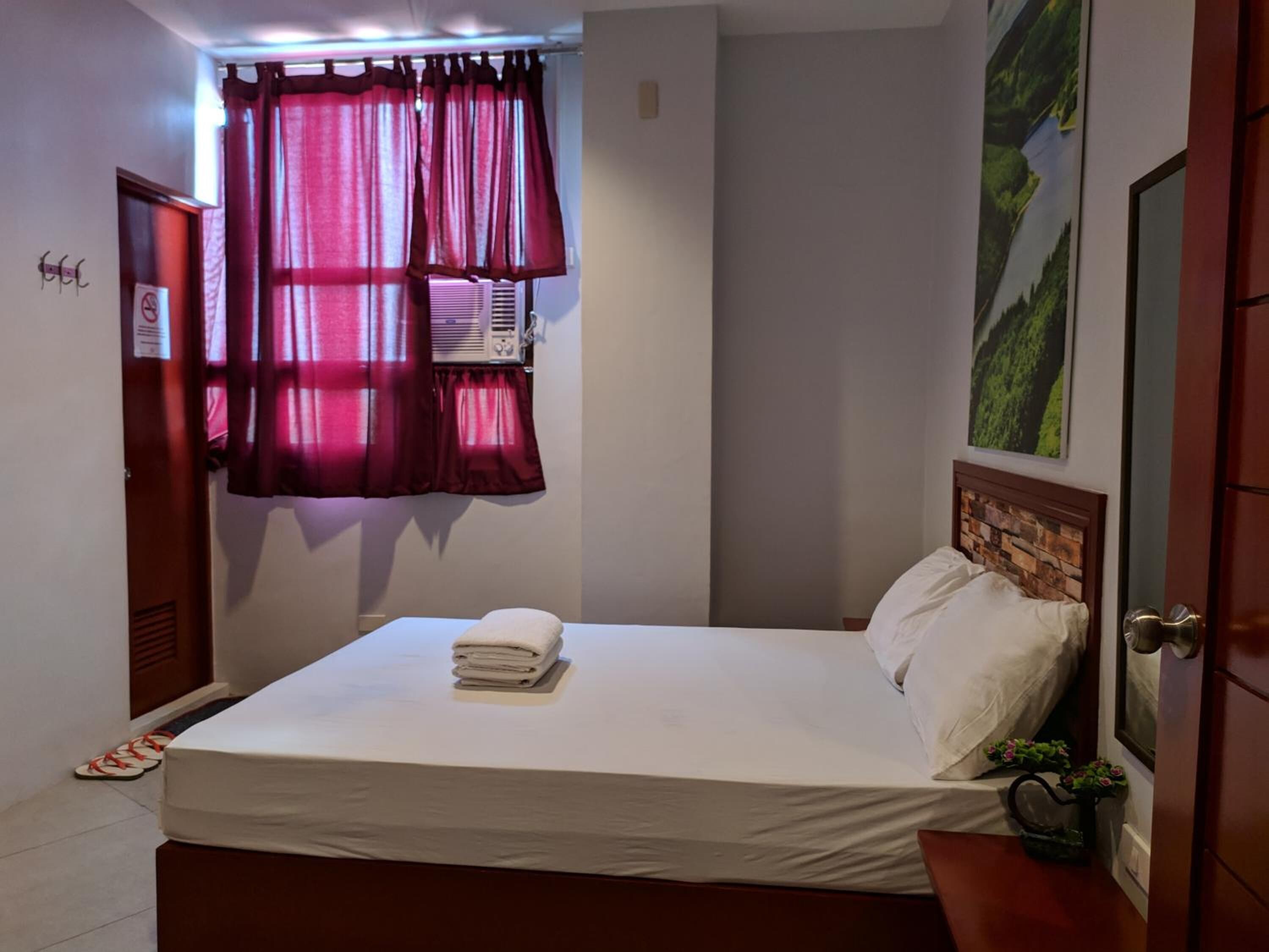 A neatly made bed with white linens and a folded towel, a window with red curtains, and a wall-mounted air conditioner.