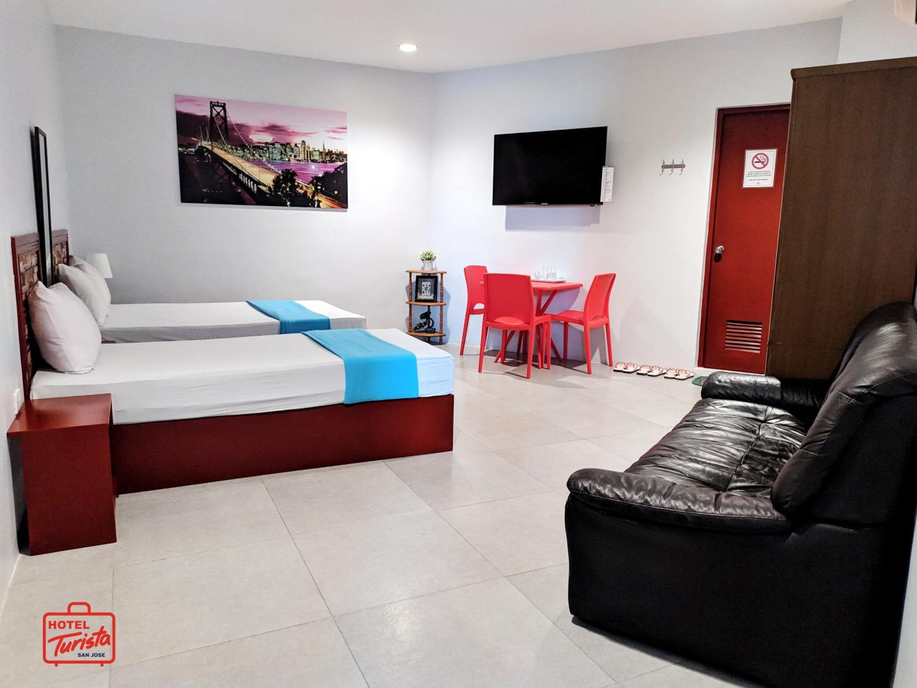 A hotel room with a bed, a couch, a TV, a red door, and a painting on the wall.