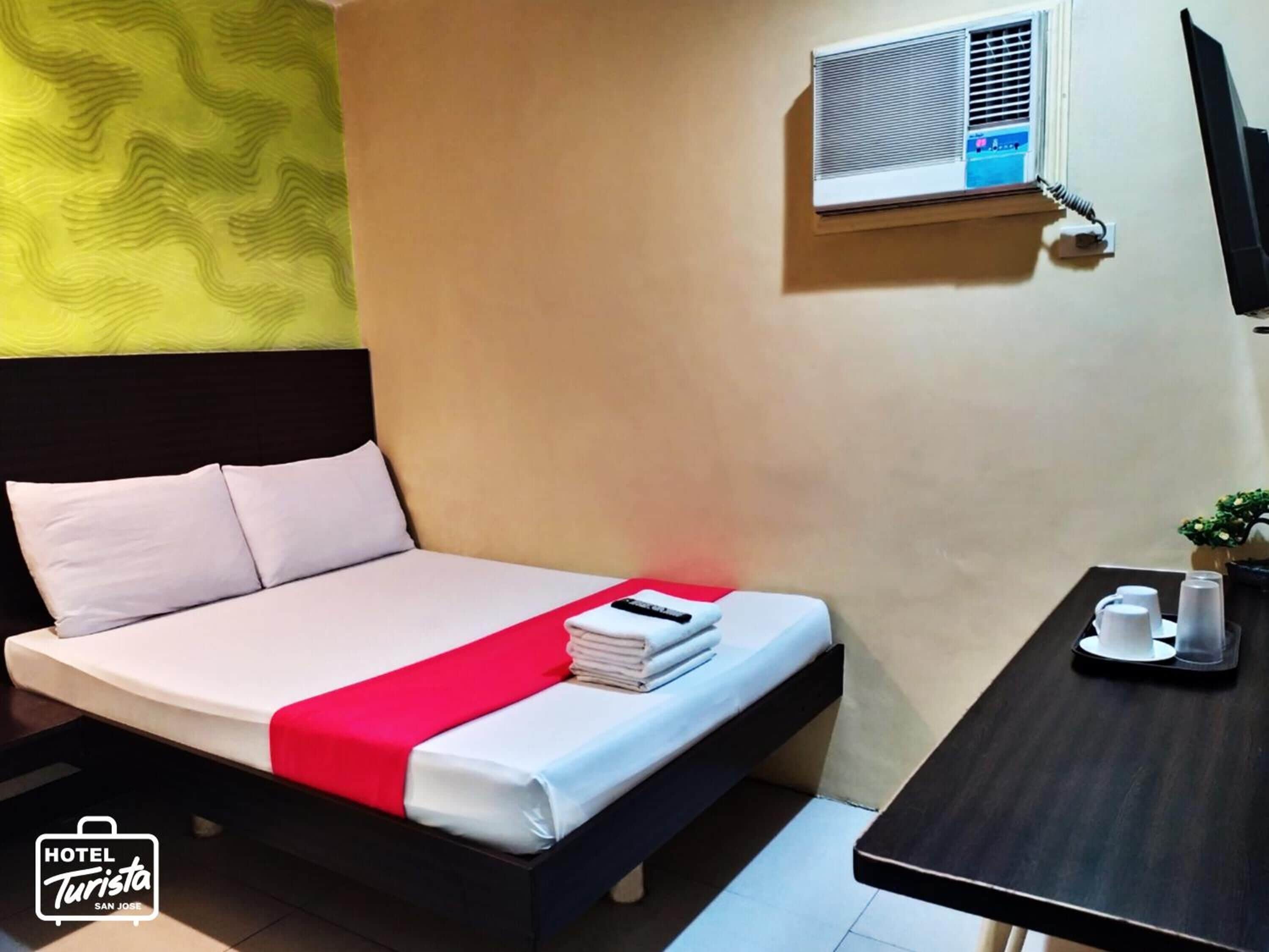 A hotel room with a bed, a small table with cups and a tray, and an air conditioning unit on the wall.
