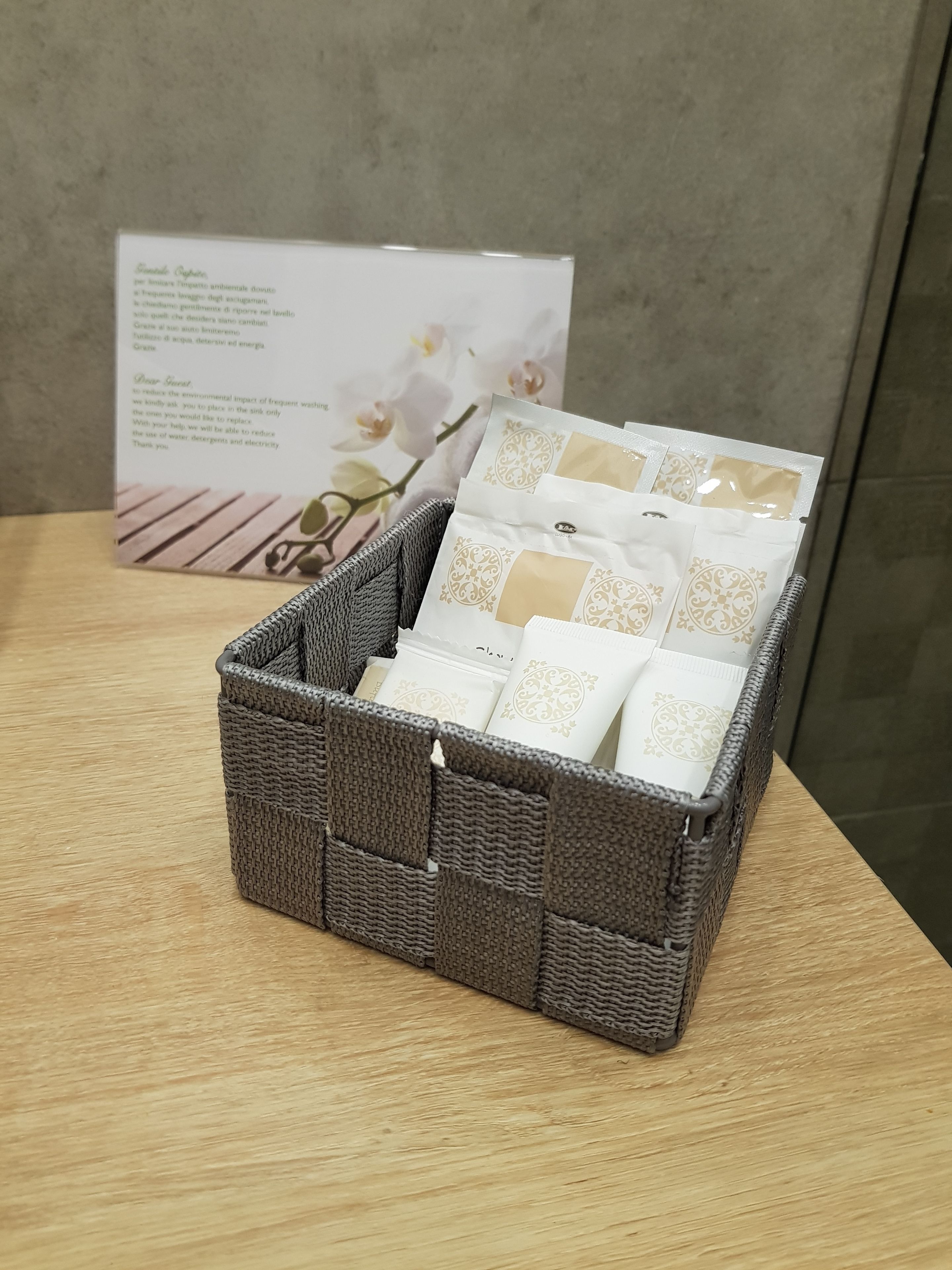 A wicker basket containing several white and beige packaged items, placed on a wooden surface with a floral-patterned card in the background.