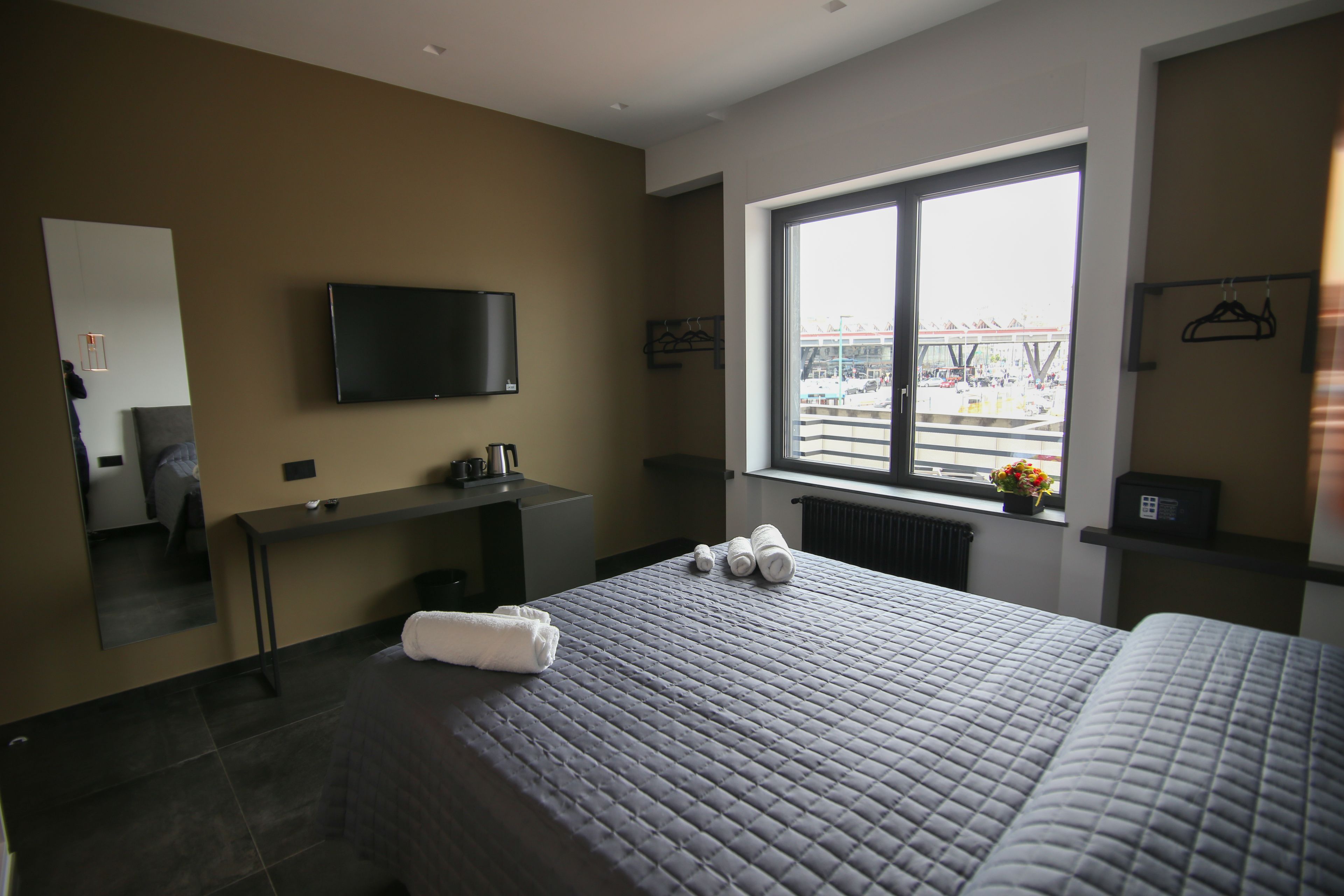 A hotel room with a bed, a desk, a TV, a window with a view of the city, and a radiator.