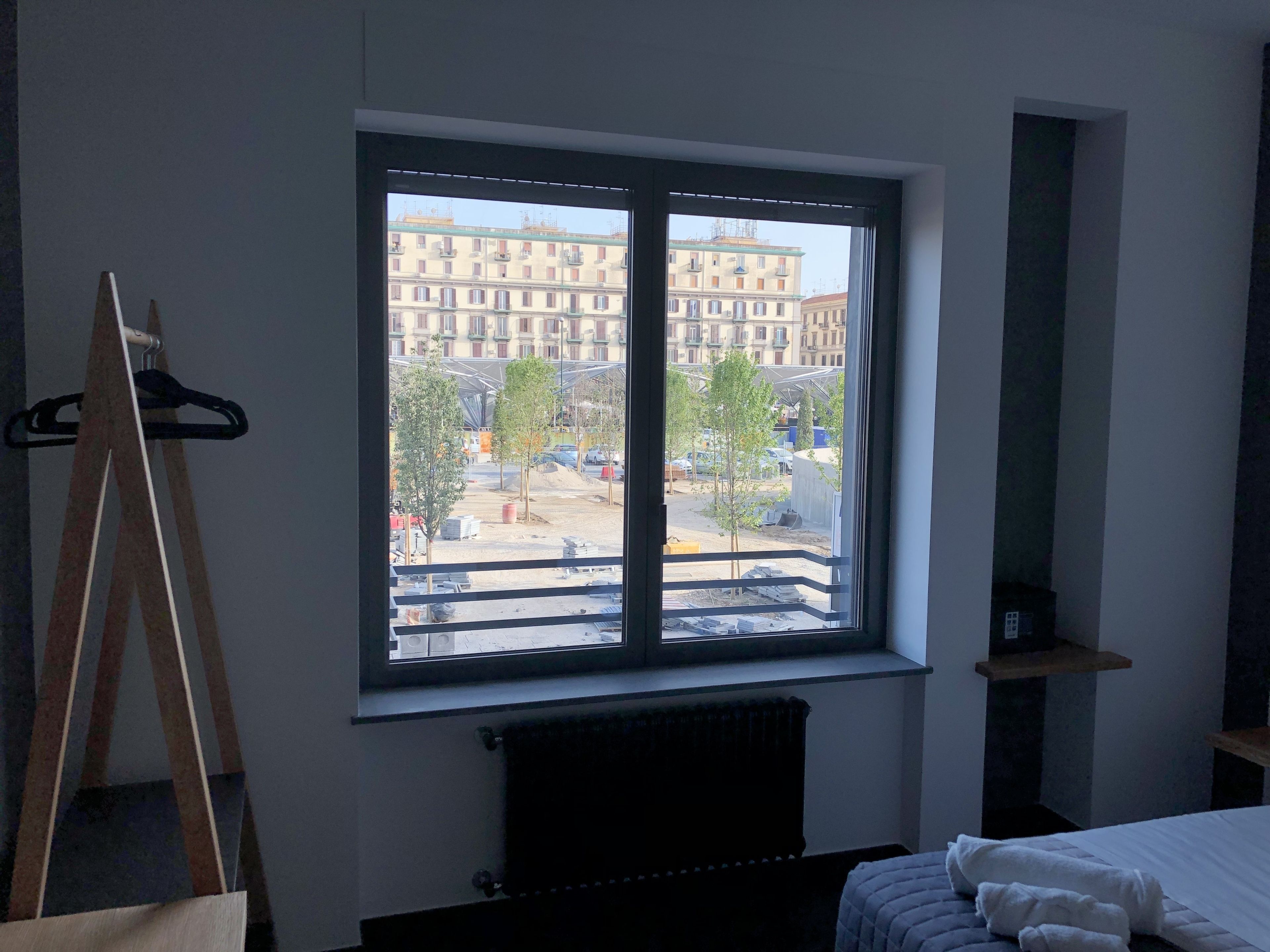 Ein Zimmer mit großem Fenster, einem Heizkörper darunter, einem Bett mit weißer Bettwäsche und einer Staffelei aus Holz links.