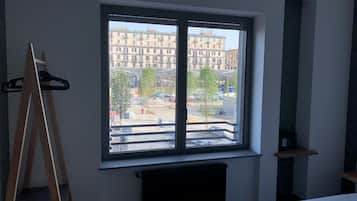 Ein Zimmer mit großem Fenster, einem Heizkörper darunter, einem Bett mit weißer Bettwäsche und einer Staffelei aus Holz links.