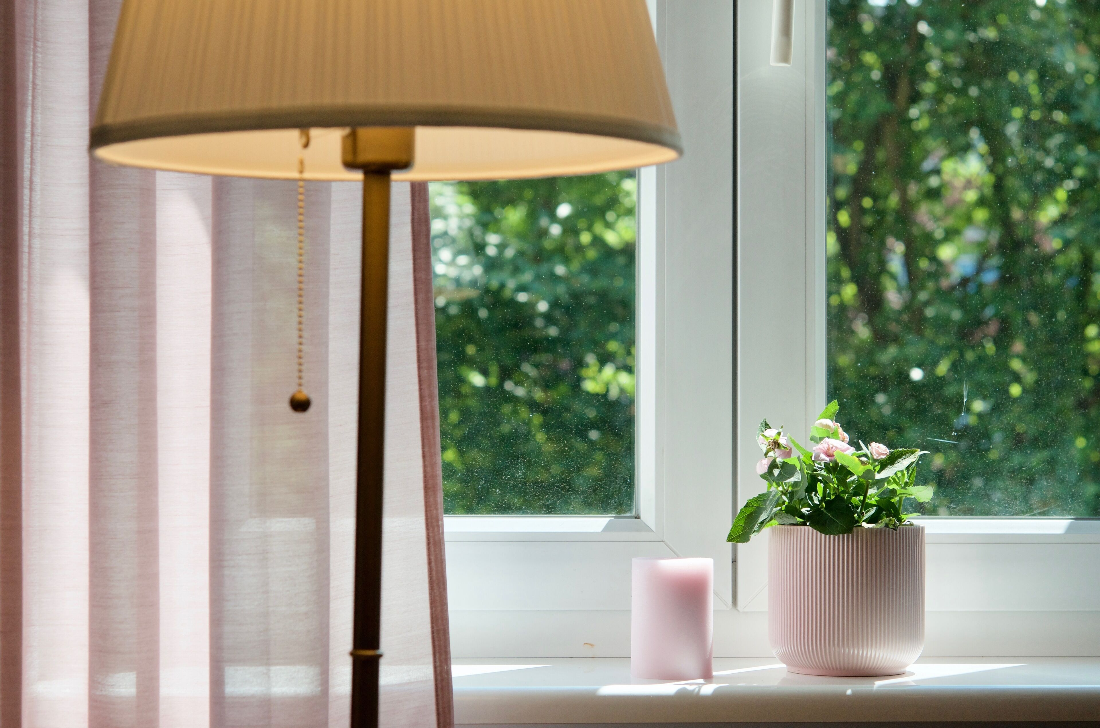 Una ventana con vistas a la vegetación, una vela rosa y una lámpara en el alféizar.