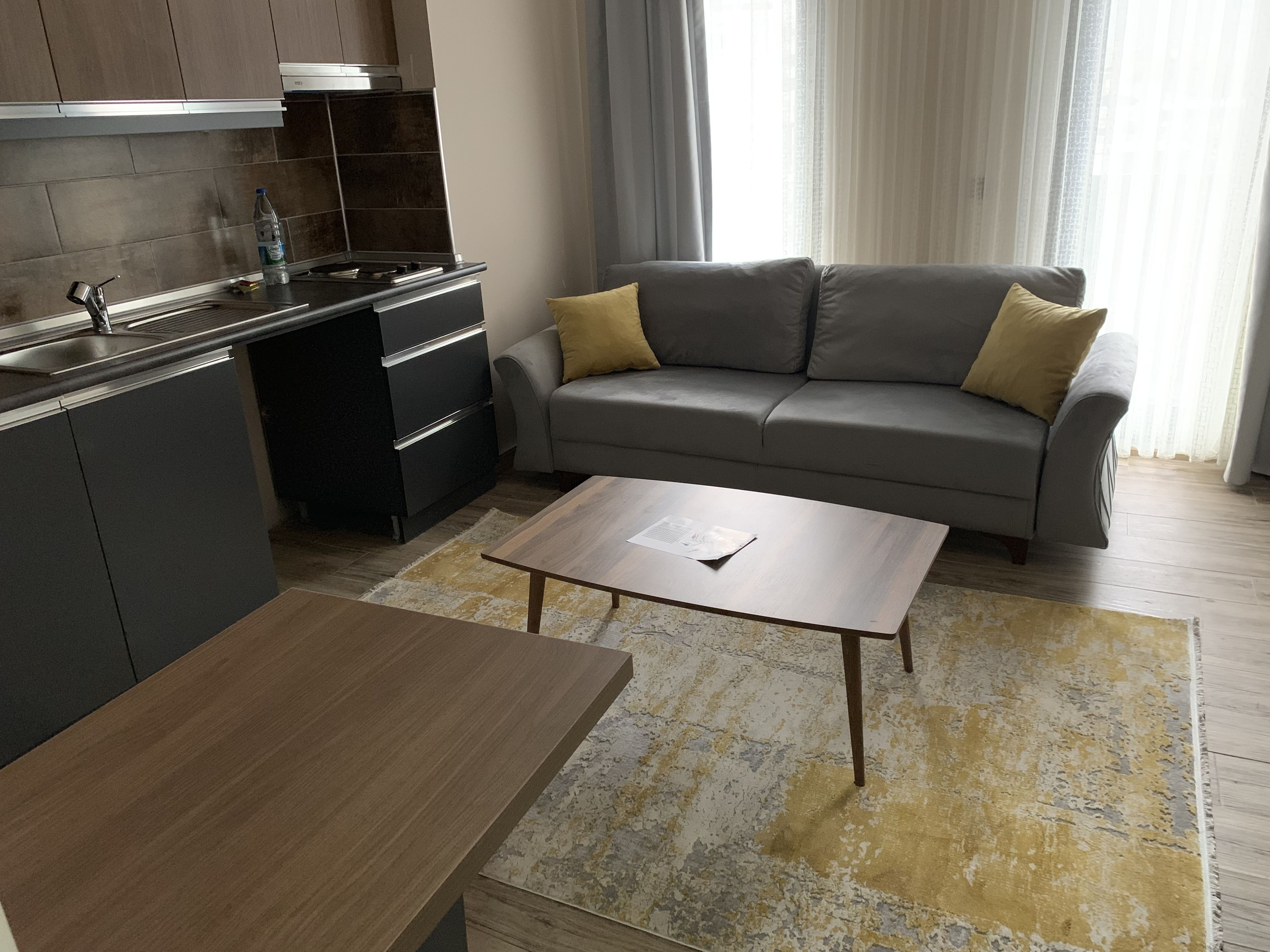 A modern kitchen with dark cabinets and a stainless steel range hood, a grey sofa with yellow cushions, and a wooden coffee table.