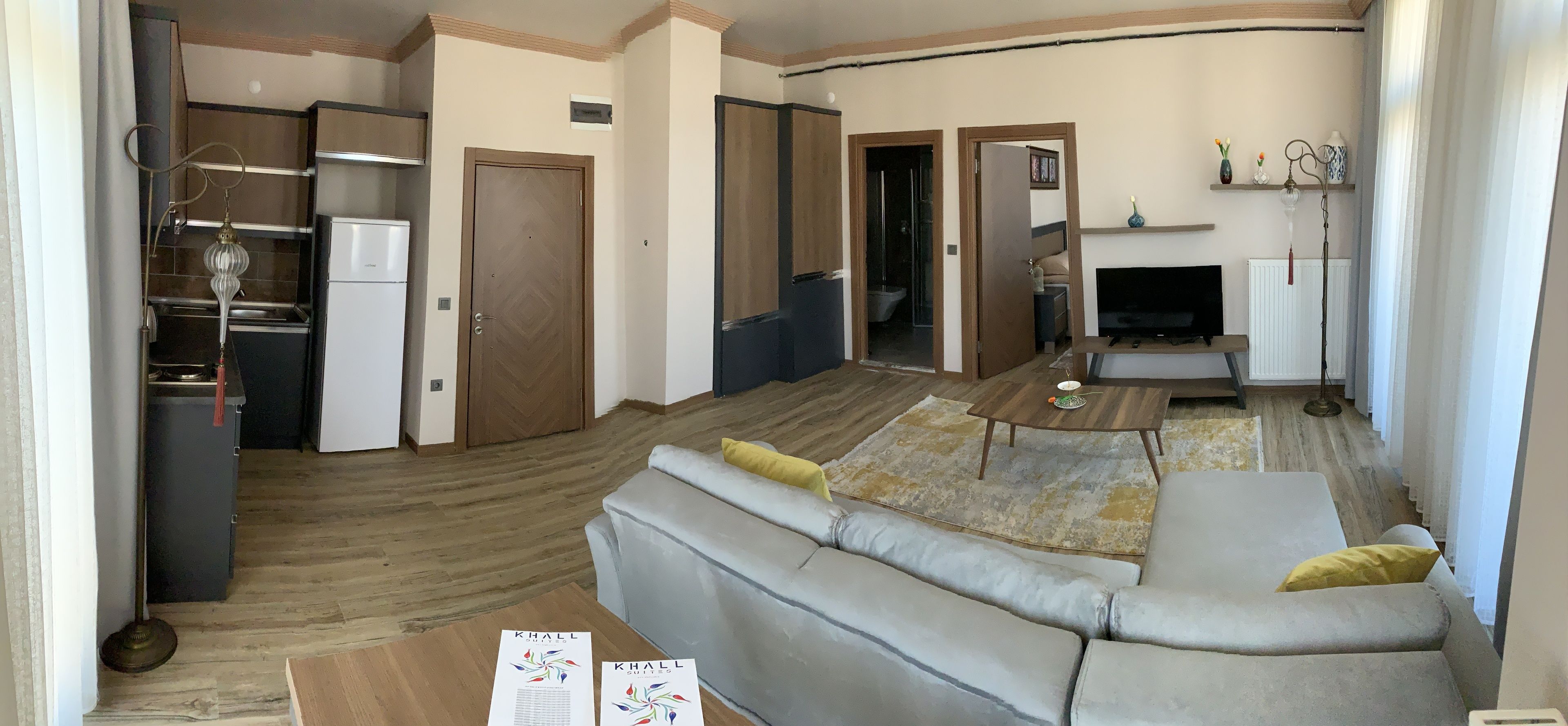 Un salon moderne avec un canapé gris, une table basse en bois et un téléviseur. On aperçoit un coin cuisine avec un réfrigérateur et des placards, ainsi qu'une salle de bain visible à travers une porte ouverte.