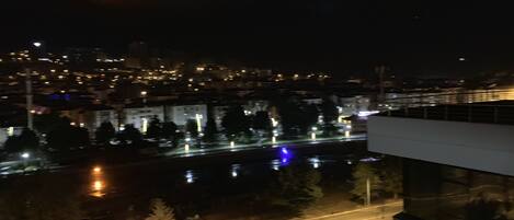 Une vue nocturne d’une ville, avec une rivière reflétant les lumières, un pont et une route bordée d’arbres.
