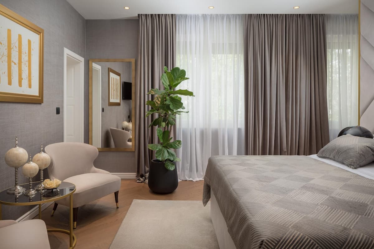 A modern hotel room with a large bed, a sitting area with a chair and a small table, a large mirror, and a potted plant.