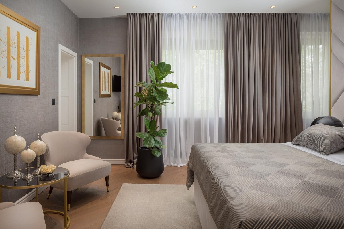 A modern hotel room with a large bed, a sitting area with a chair and a small table, a large mirror, and a potted plant.