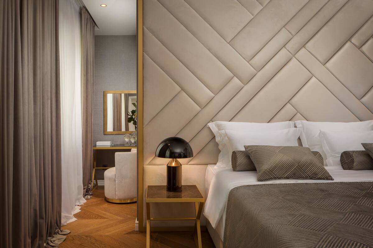 A modern hotel room with a large bed, a nightstand with a lamp, a wooden floor, and a large mirror on the wall.