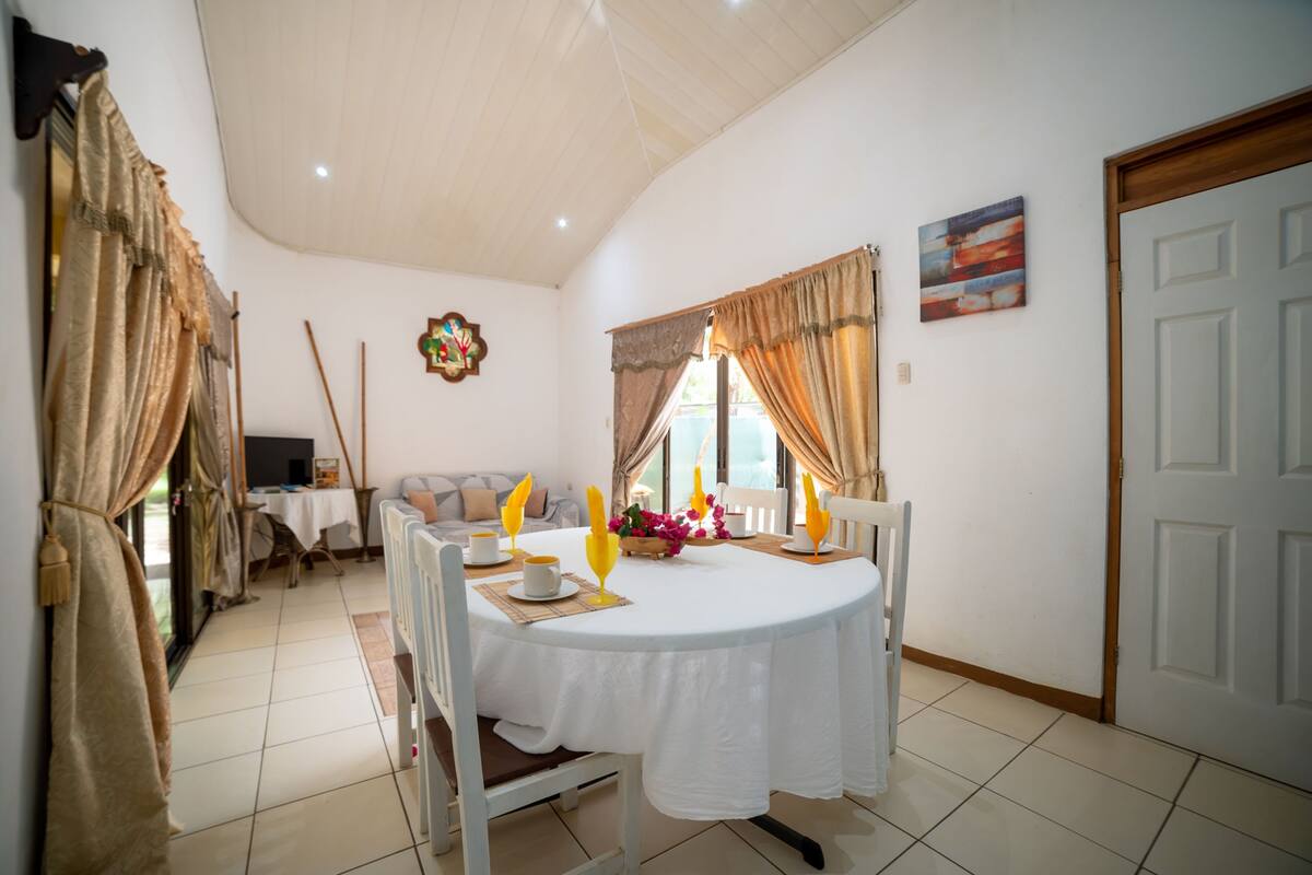 A dining area with a round table, chairs, and a white tablecloth. There is a sofa and a television in the background.
