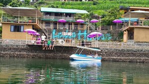 A waterfront property with a stone wall, a small boat, and a building with purple umbrellas. - The Lake House (Pokhara)