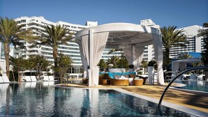 Zona de piscina con un área de descanso cubierta, sillas de descanso y vista despejada de un moderno edificio hotelero al fondo.