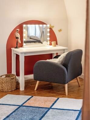 A room with a vanity table, a chair, a mirror with lights, a candle, and a decorative item.