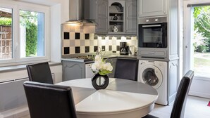 A modern kitchen with grey cabinetry, a built-in oven, a washing machine, and a dining area with a round table and chairs. - Gite La Tour - Cocoon for 2 with pool and nature (Saint-Geyrac)