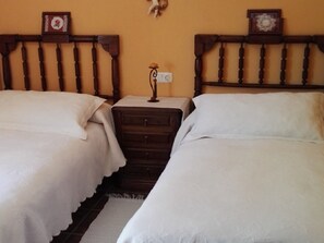 A room with two single beds, a wooden headboard, a nightstand with a lamp, and a wall clock. - El Condado Casa Rural (Fuentes de Onoro)