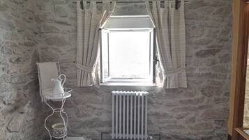 A room with a stone wall, a radiator, a window with curtains, and a small table with a pitcher and cups.