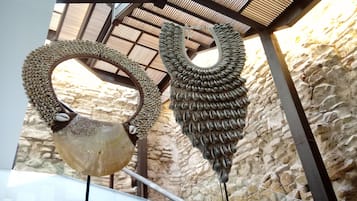 Una escultura con base circular y estructura semejante a un ala, exhibida en interiores frente a un fondo de pared de piedra y un techo con marco metálico.