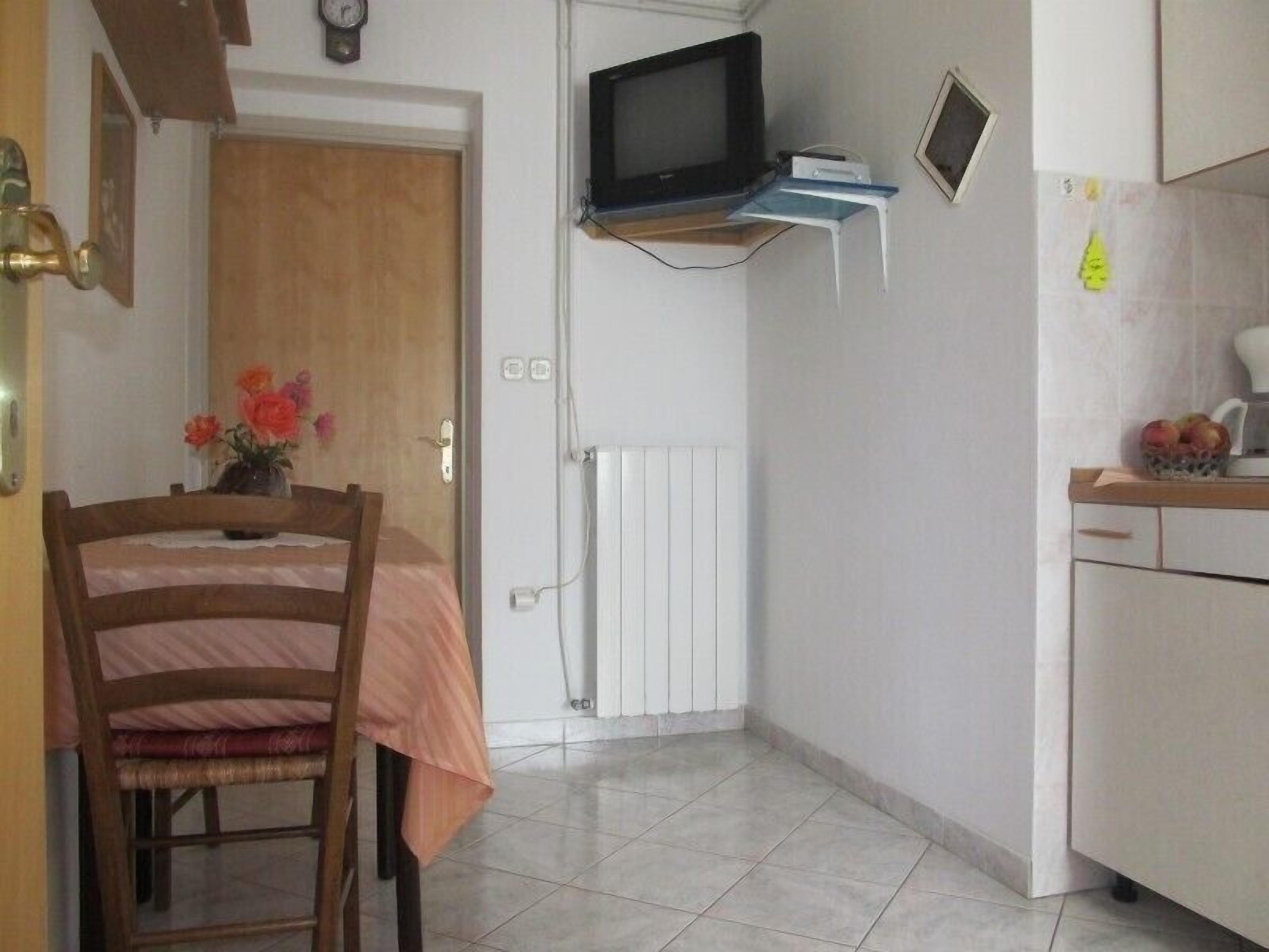 A small, compact kitchen with a microwave, a small TV, and a refrigerator. There is a wooden chair and a table with a pink tablecloth. A small vase with flowers is on the table.