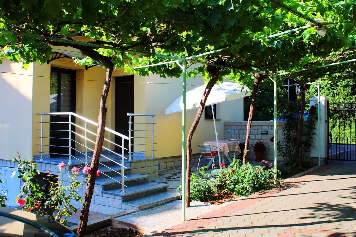 A residential building with a staircase leading to a door, a small garden area with plants and flowers, and a metal railing.