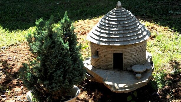 A small, stone birdhouse with a conical roof, placed on a circular stone base, surrounded by grass and a small tree. - House Marija (Pula)