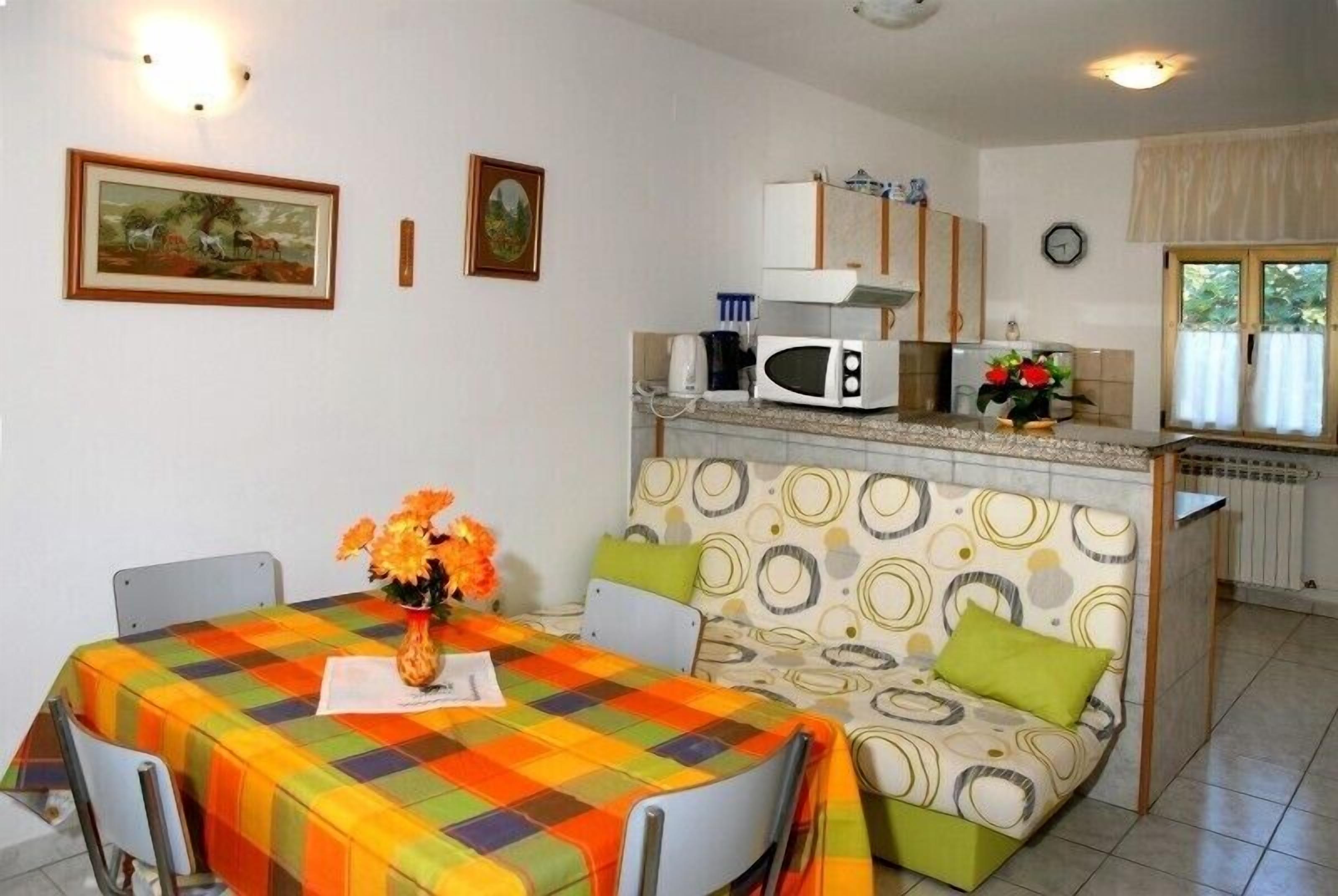 A compact kitchen with a microwave, a small refrigerator, and a sink. There is a dining table with a colorful tablecloth and a vase of flowers. The room has a patterned sofa and a window with curtains.