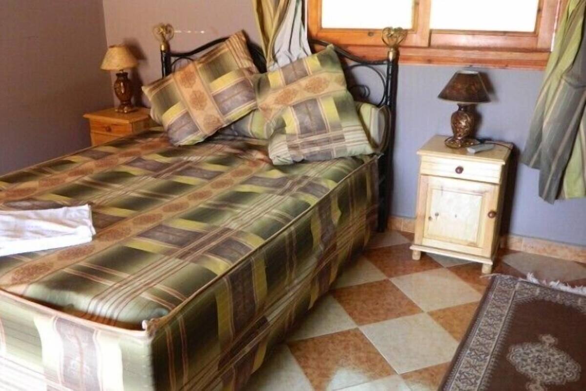Een bed met een geruite sprei, twee kussens, een nachtkastje met een lamp en een kleed op de vloer.