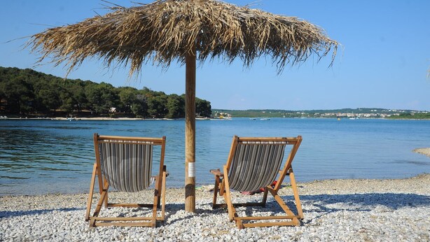 Two wooden beach chairs under a thatched umbrella on a pebbled beach with a calm sea and a distant shoreline. - Villa Nino (Liznjan)