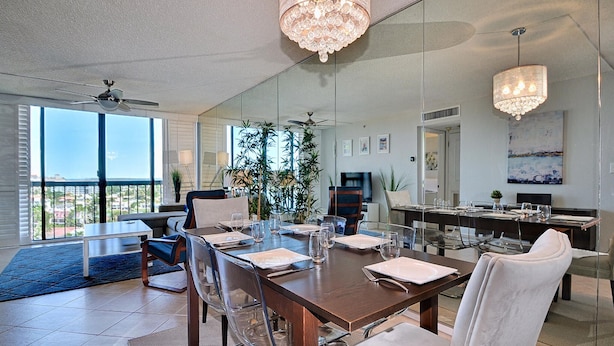 A modern dining room with a large table set for six, surrounded by clear chairs. A large window offers a view of the cityscape. - Dolphin Cove  PH 9 '90 day min stay with 2 week lead time' (Clearwater Beach)
