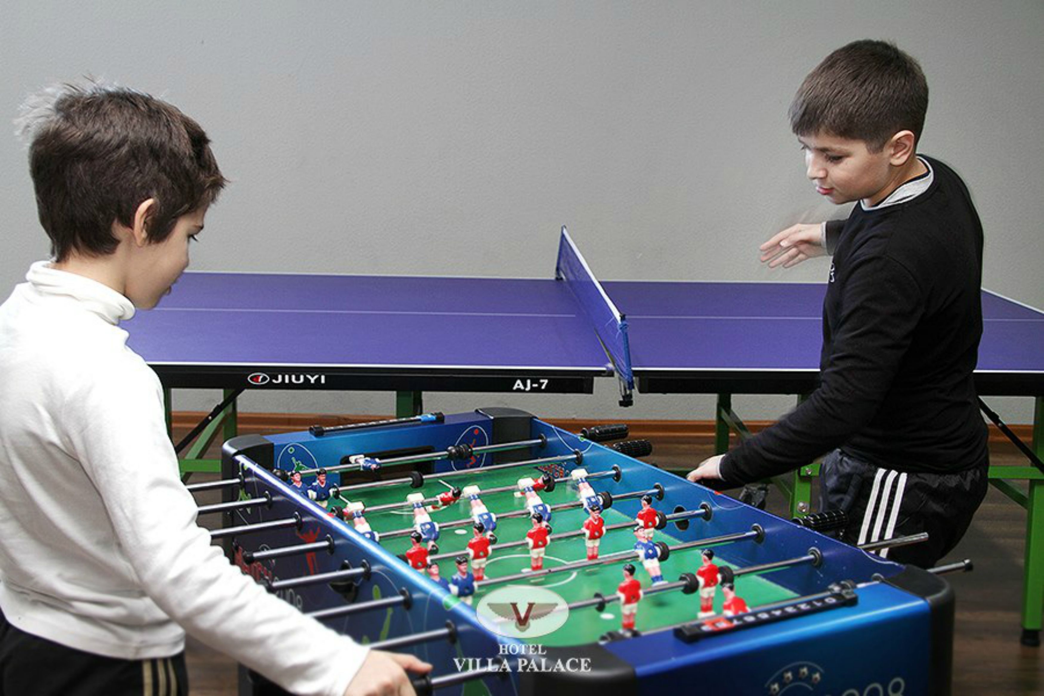 Duas pessoas jogando pebolim numa sala com uma mesa roxa e um logotipo visível na mesa.