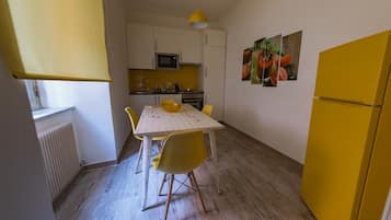 Een compacte keuken met witte kasten, een magnetron en een gele koelkast. Er staat een eettafel met gele stoelen en een gele hanglamp. Aan de muur hangt een schilderij met fruitmotieven.