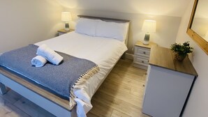 Een net opgemaakt bed met witte lakens en een blauw-wit gestreept deken, twee nachtkastjes met lampen, een klein kastje en een potplant op een houten dressoir.
