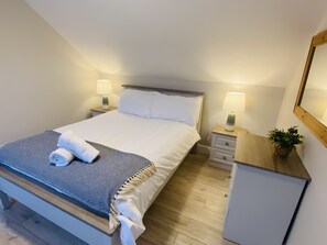 A neatly made bed with white linens and a blue and white patterned throw, two bedside tables with lamps, a small cabinet, and a potted plant on a wooden dresser.