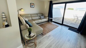 Een moderne woonkamer met een bank, een klein bijzettafeltje, een lamp en uitzicht door een grote schuifdeur op een buitenruimte.
