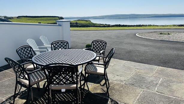 Een ronde tafel met zes stoelen, een witte muur en uitzicht op zee en heuvels.