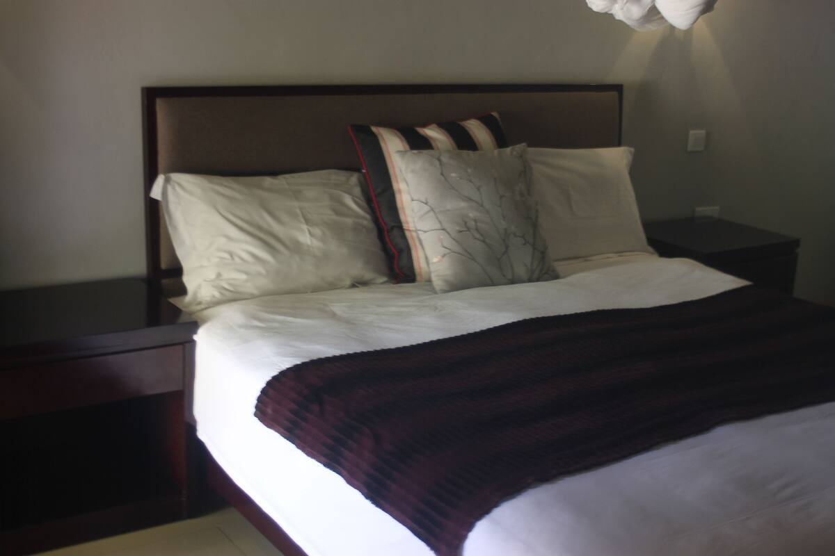 Een net opgemaakt bed met witte lakens en een donkerbruine plaid. Er liggen twee kussens op, waarvan één met een ontwerp van een tak en de ander effen wit. Het hoofdbord is van donker hout, en er staat een klein houten nachtkastje naast het bed.