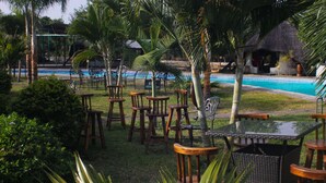 Un área junto a la piscina con sillas y mesas de madera, rodeada de palmeras y una estructura con techo de paja.