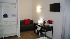 Ein gemütlicher Raum mit Schreibtisch, Stuhl, Sofa, Fernseher, Kleiderschrank und einer kleinen Pflanze.