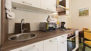 A modern kitchen with white cabinets, a stainless steel sink, a microwave, a coffee maker, and a dining area with a yellow bowl of fruit. - Apartman Snježana (Pula)