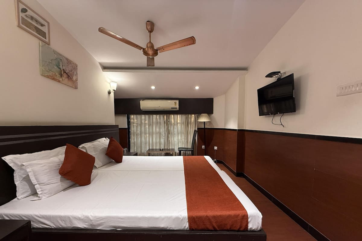 A hotel room with a large bed, a ceiling fan, an air conditioning unit, a TV mounted on the wall, and a window with curtains.