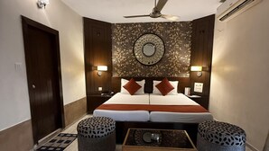 A bedroom with a bed, two ottomans, a coffee table, a ceiling fan, and wall-mounted lights. - Palm Village Resort (Alipur)