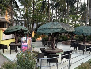 An outdoor seating area with tables and chairs under tree-shaped umbrellas, surrounded by palm trees and a building with a yellow facade. - Palm Village Resort (Alipur)