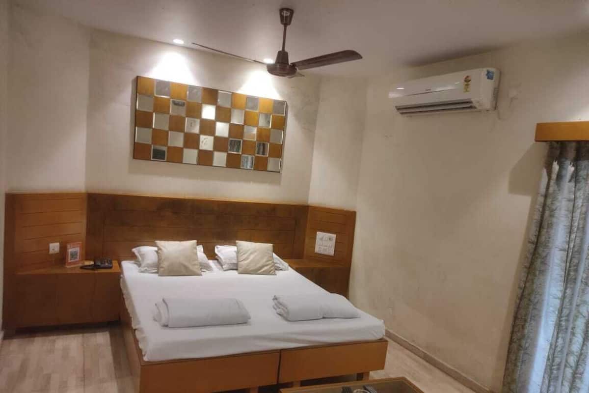 A hotel room with a double bed, wooden headboard, a small bedside table, a glass coffee table, an air conditioning unit, and a ceiling fan.