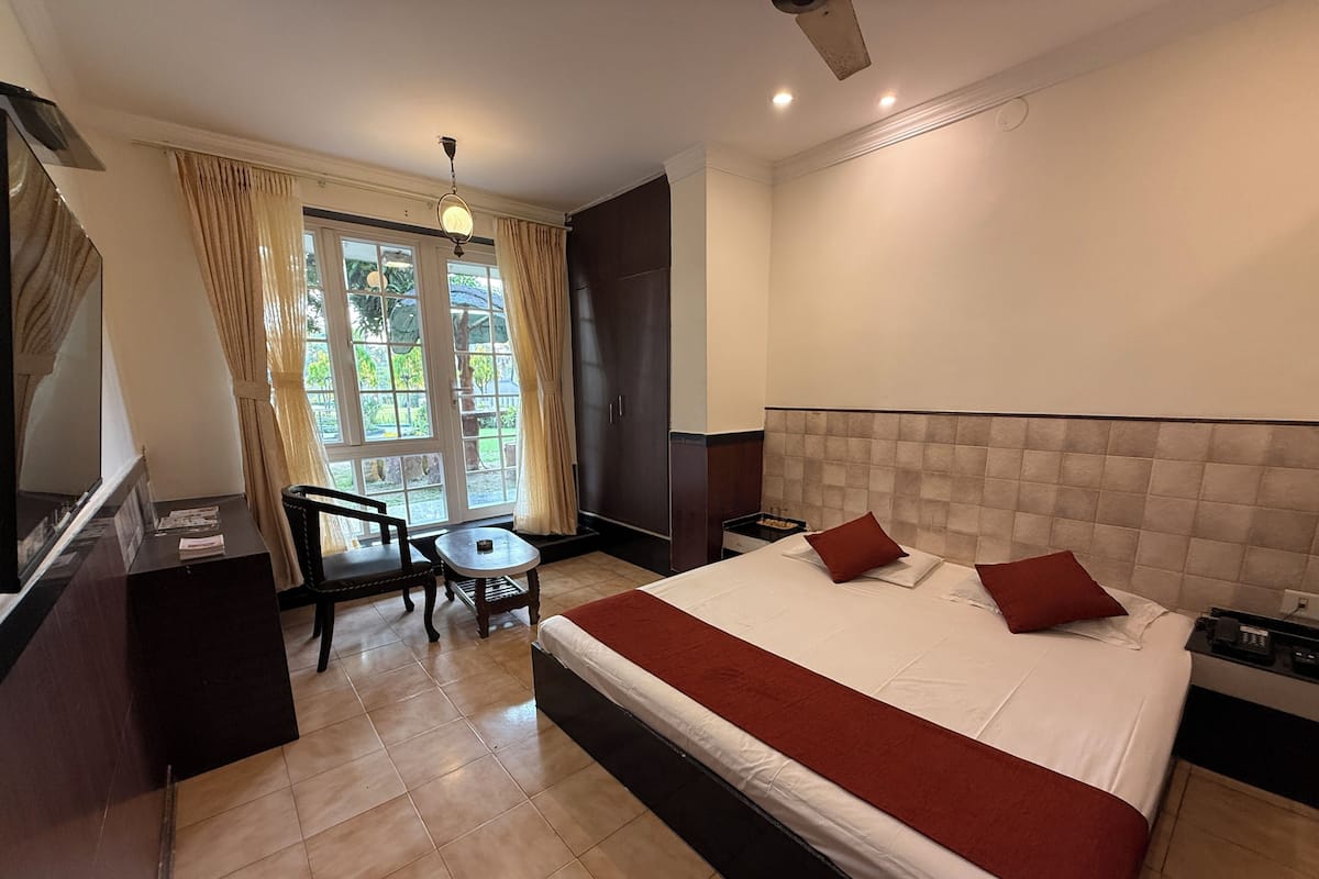 A hotel room with a large bed, a desk, a chair, and a view of greenery through the window.