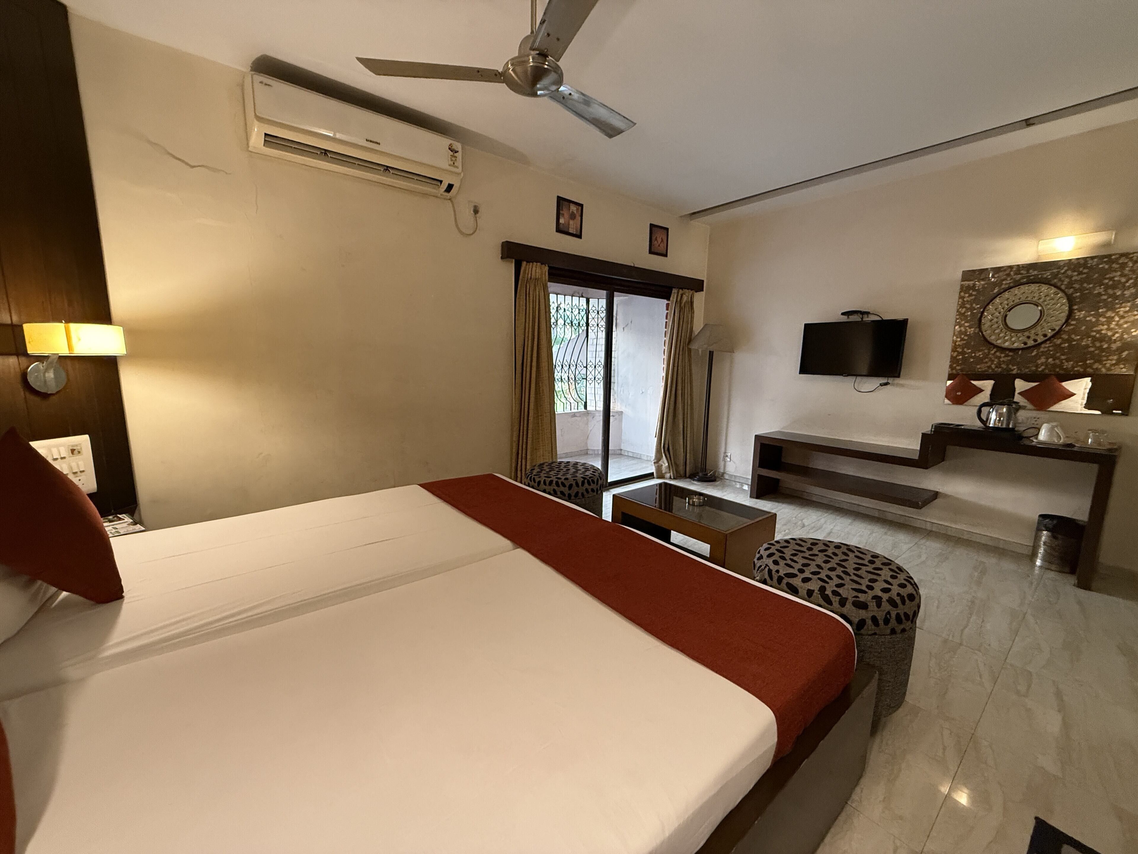 Habitación de hotel con una cama grande, ventilador de techo, aire acondicionado, televisor, una mesita pequeña, un banco y una ventana con cortinas.