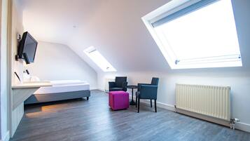 A modern room with a bed, a pink ottoman, two chairs, a small table, and a TV mounted on the wall.