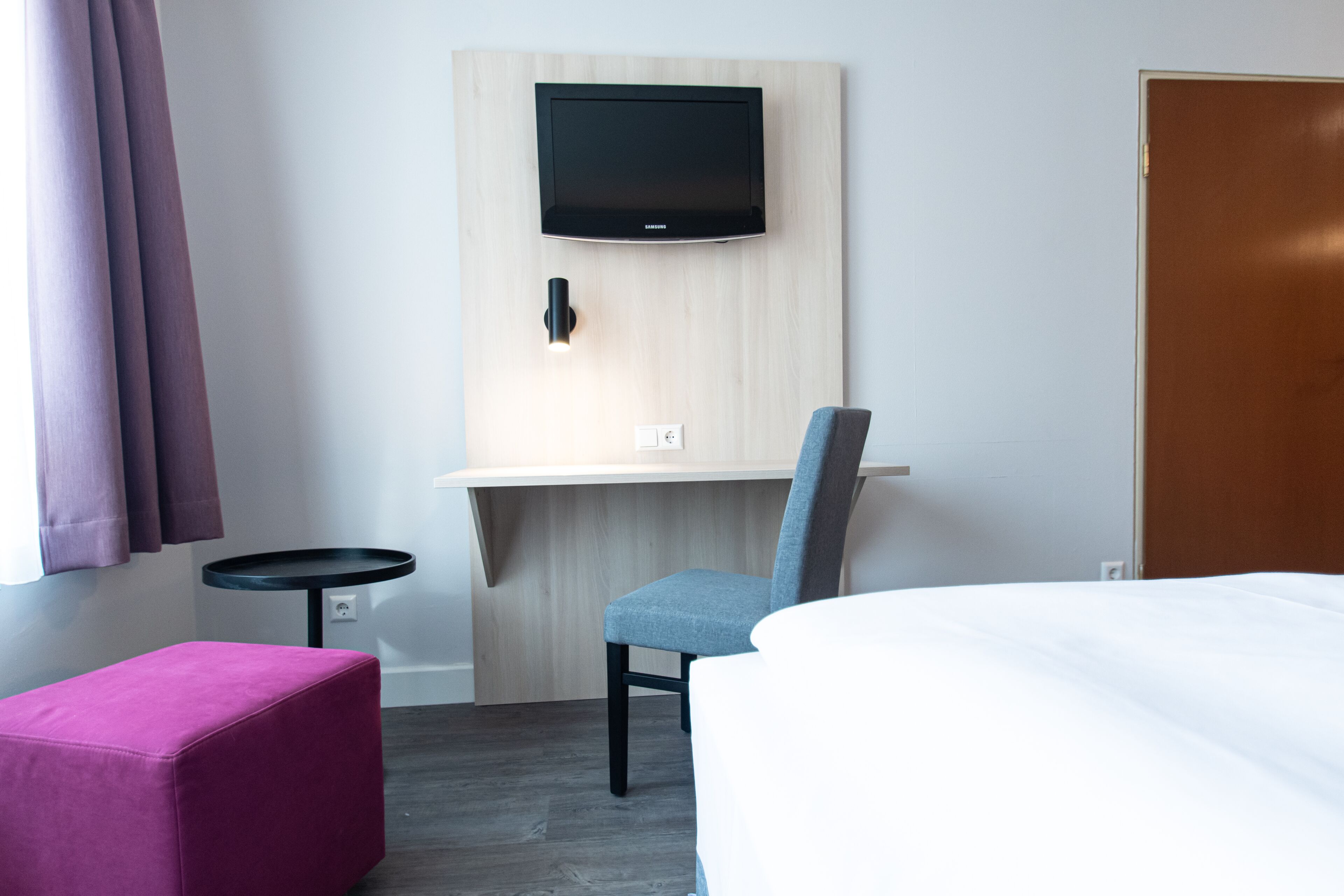 A hotel room with a bed, a desk with a chair, a television mounted on the wall, and a small round table.