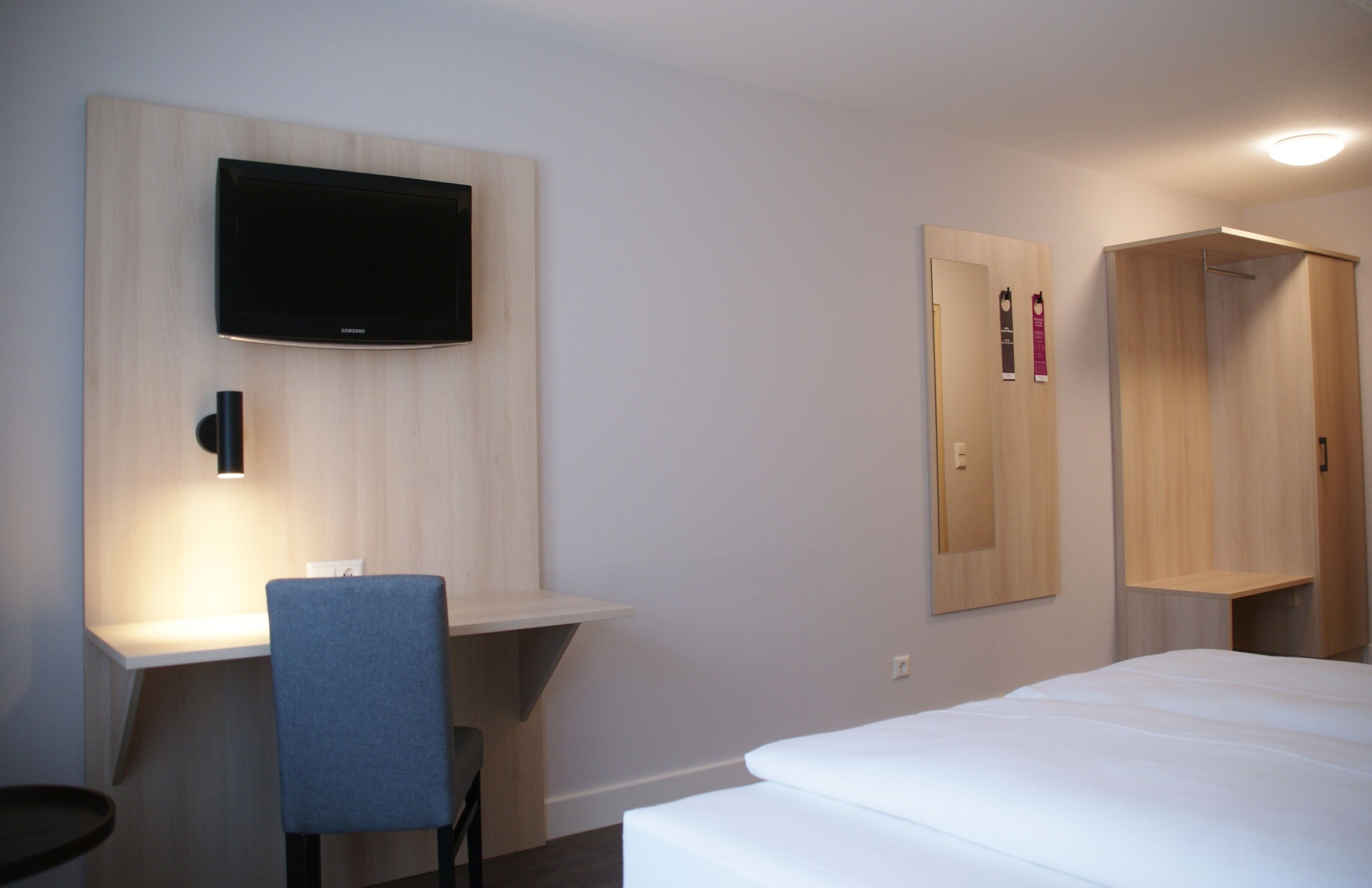 A hotel room with a bed, a desk with a chair, a flat-screen TV mounted on the wall, and a wardrobe.